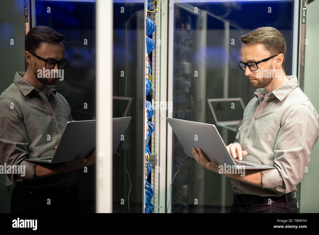 Serious busy young IT engineer with beard standing by server cabinet and using laptop while providing network support Stock Photo