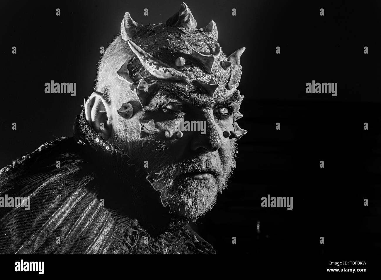 Demon with golden collar on black background. Man with thorns or warts, face covered with glitters. Senior man with white beard dressed like monster Stock Photo