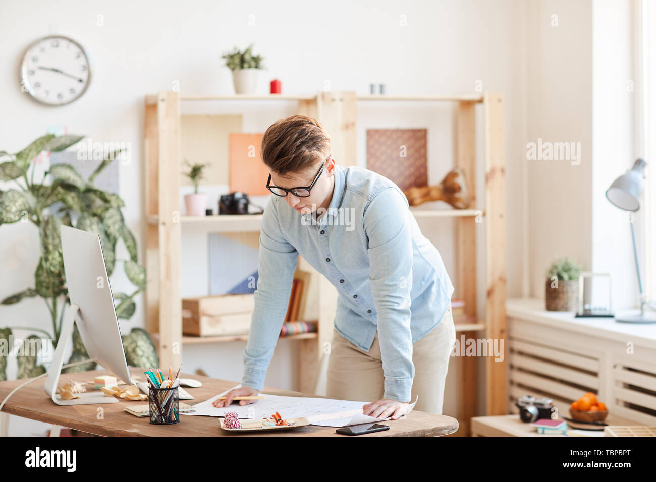 Serious pensive handsome young male engineer in glasses standing by table with modern computer and examining blueprint in office Stock Photo