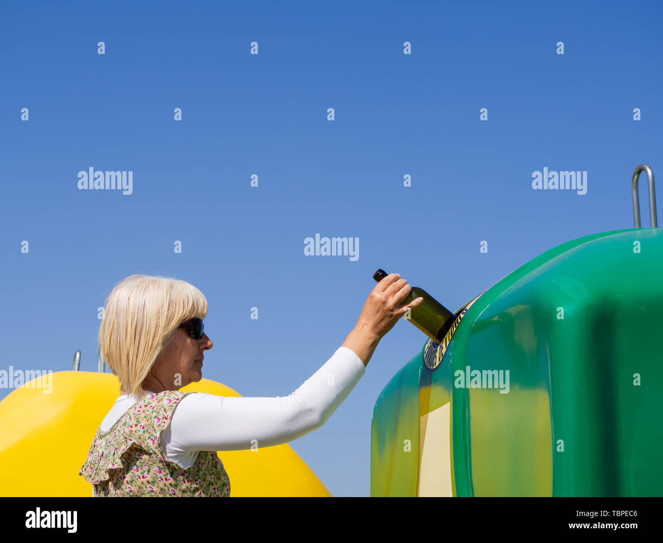 A mature woman pulling a crystal bottle in a green bin for recycling glass with the spanish text 'only glass containers' Stock Photo