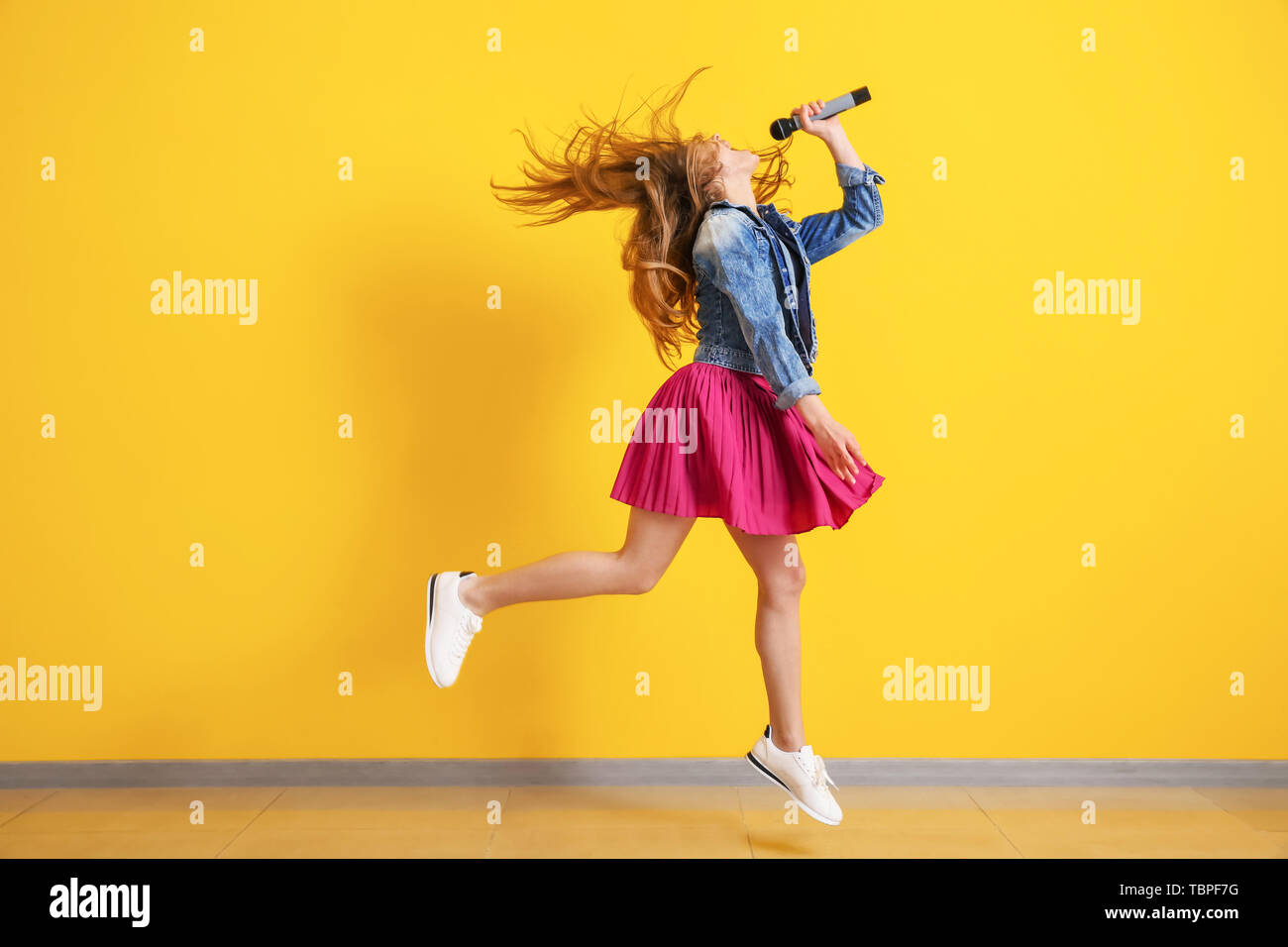 Teenage girl with microphone jumping and singing against color wall