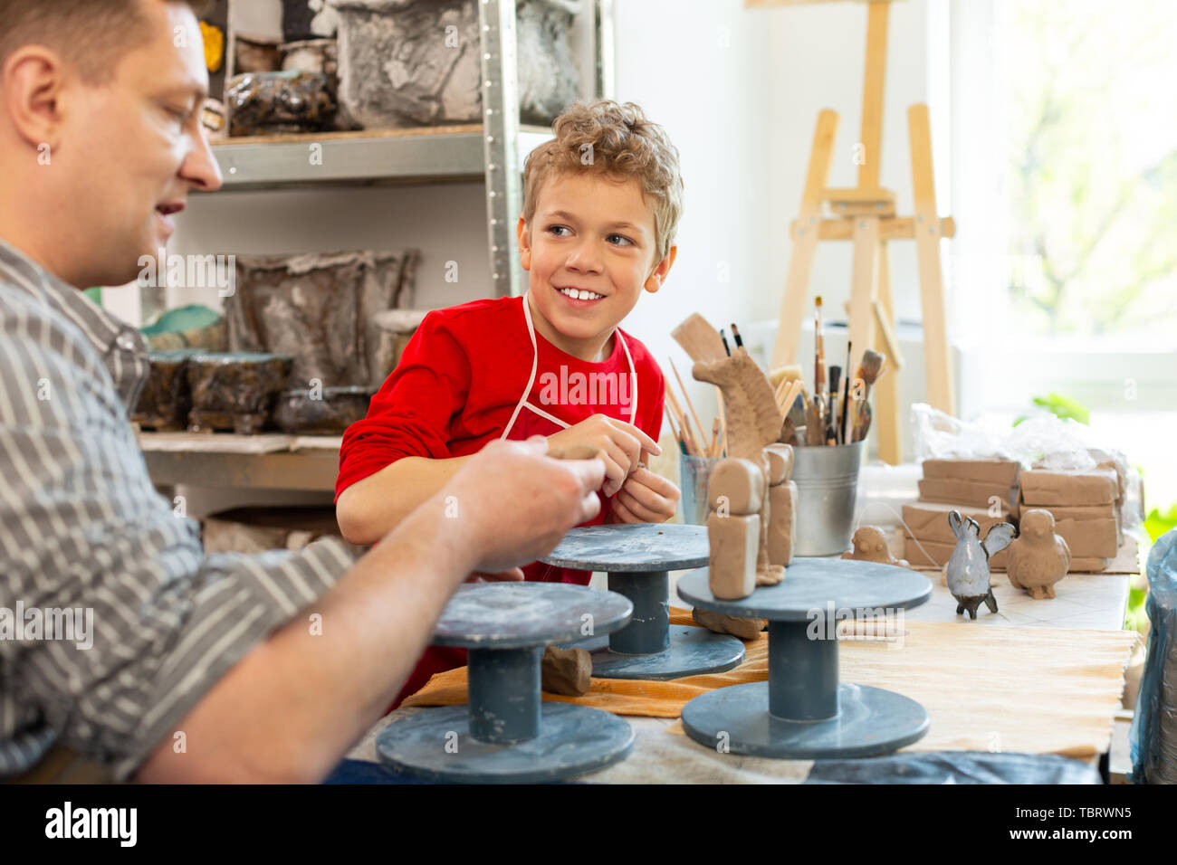 Curly boy smiling. Cheerful curly boy smiling broadly while sculpting clay animals with teacher