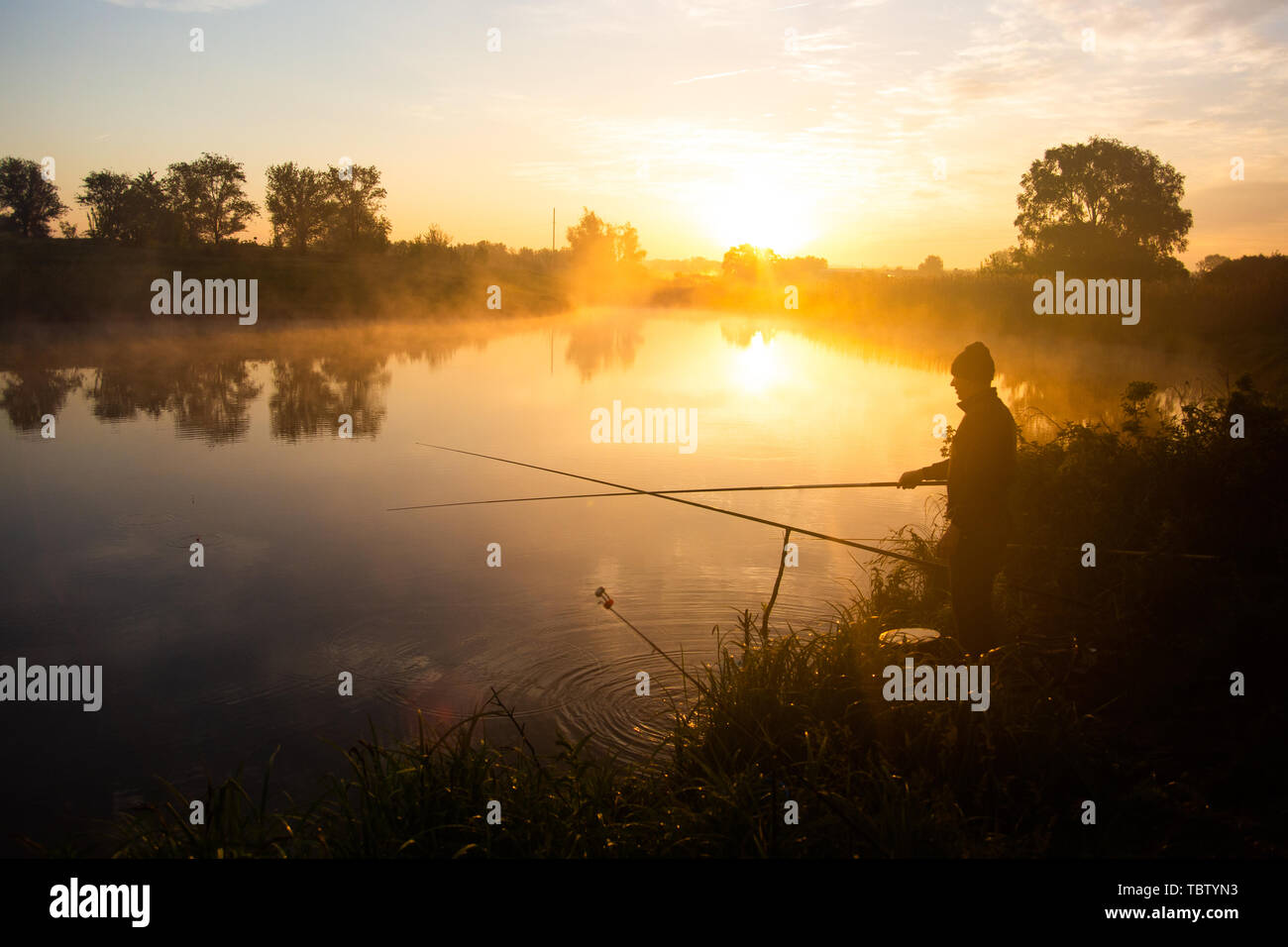 Lonely fisherman angling at foggy lake in the early morning just after golden sunrise. Stock Photo