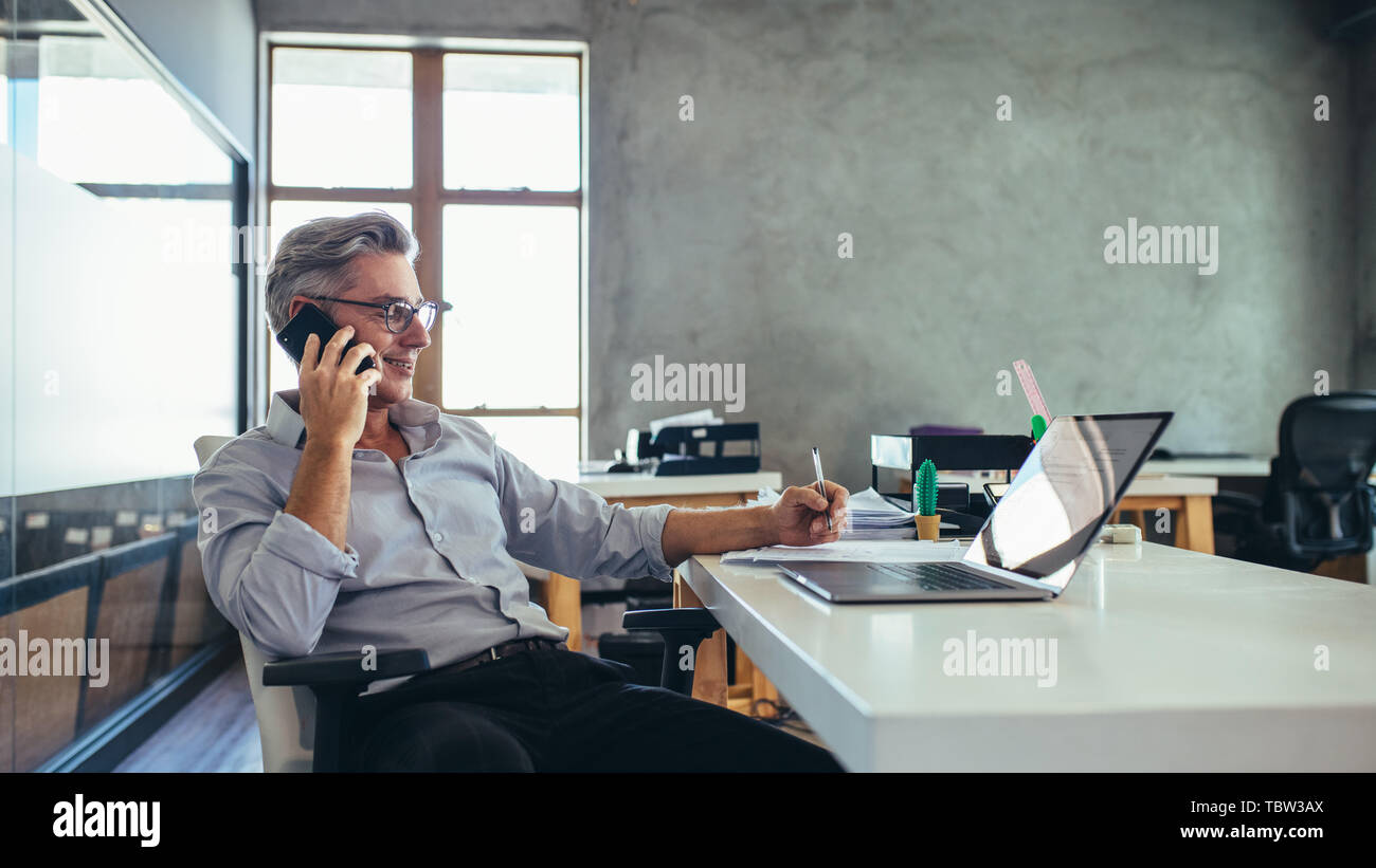 businessman on the phone sitting at the laptop in his office. Male business professional in office talking on cell phone. Stock Photo