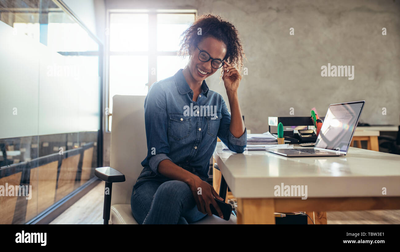 Smiling woman entrepreneur at her desk in office. Woman sitting in front of laptop in office looking at camera. Stock Photo