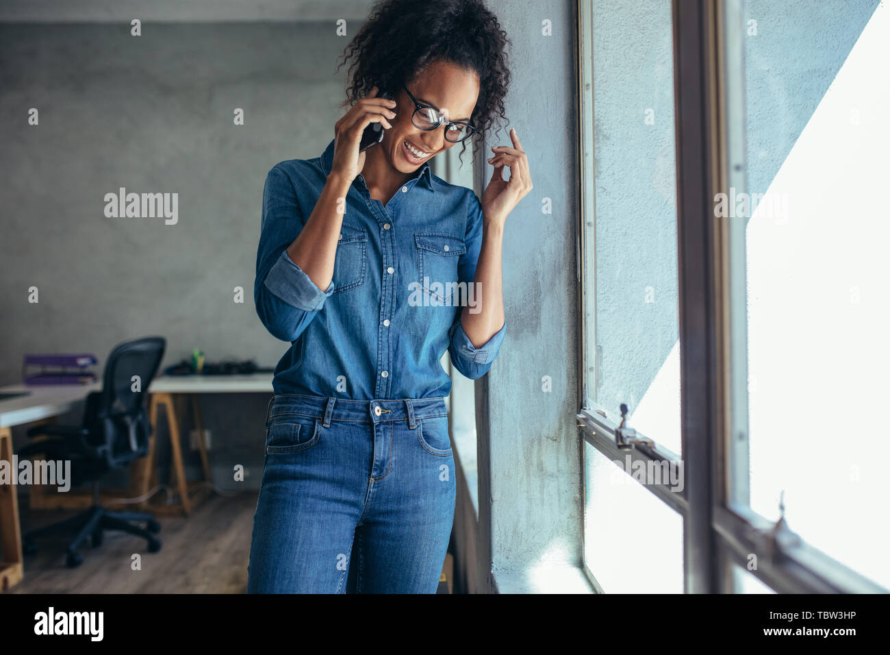 Businesswoman talking on mobile phone and smiling. Female entrepreneur standing by a window and talking over cell phone in office Stock Photo
