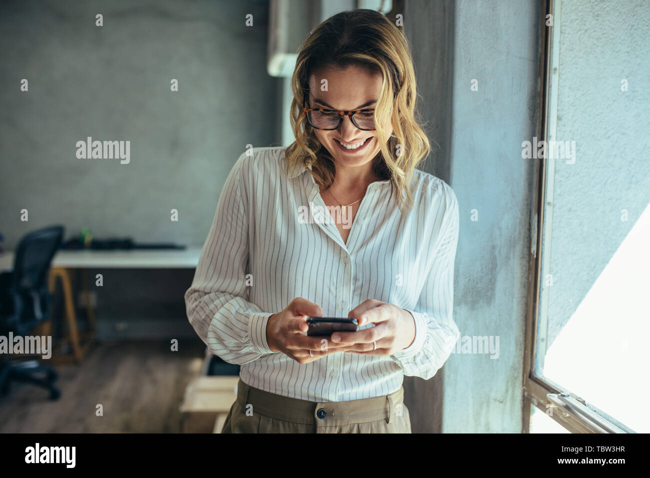 Businesswoman using her smart phone in office. Female entrepreneur looking at her mobile phone and smiling. Reading text messages. Stock Photo