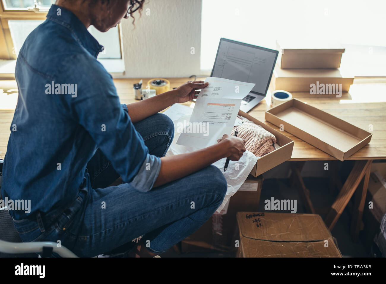 Female entrepreneur reading and verifying the invoice before shipping the product to the customer. Woman preparing shipment for delivery at her desk. Stock Photo