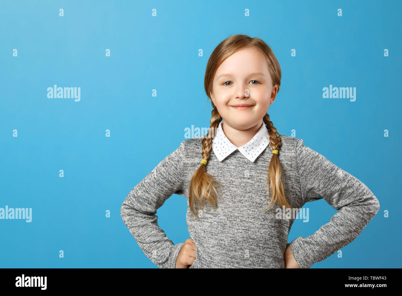 Portrait of a cute little girl on a blue background. Child schoolgirl made hands to the side and looks into the camera. Copy space. Stock Photo