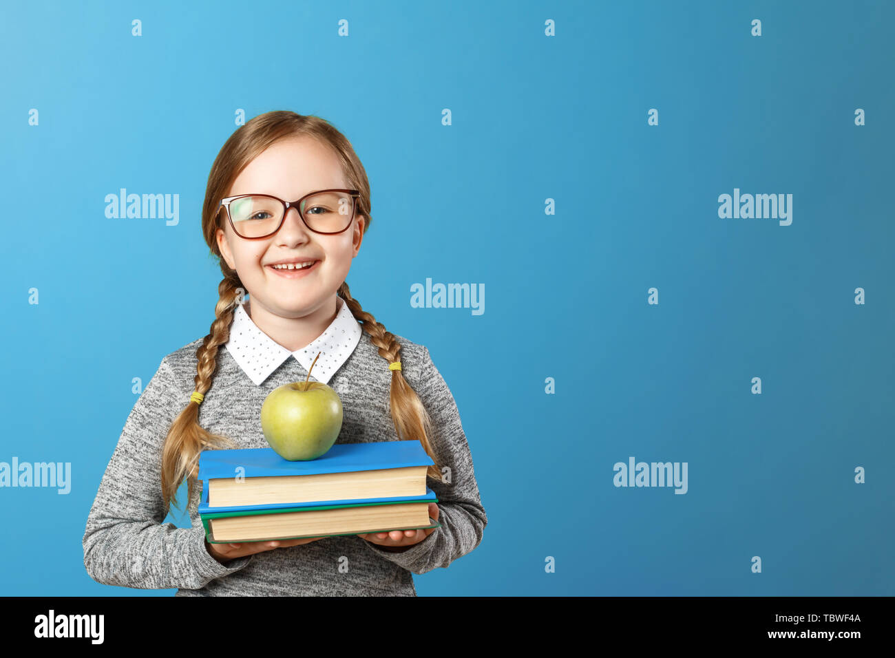 Portrait of a cheerful little girl with glasses on a blue background. Schoolgirl is holding a stack of books and an apple. Back to school. The concept Stock Photo