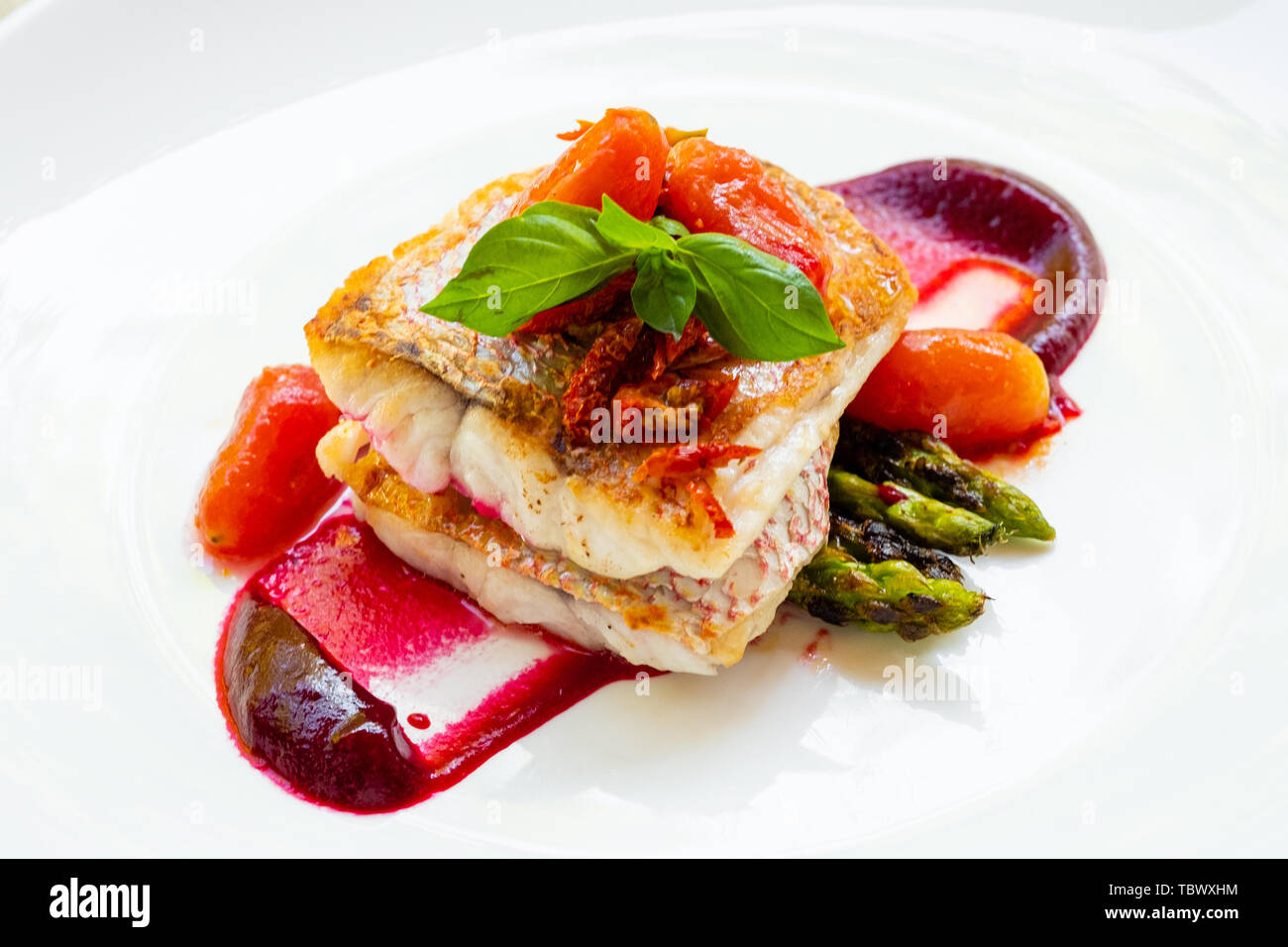 close up of a pan seared grilled red snapper fish fillet with asparagus, beetroot sauce,cherry san marzano tomatoes and basil on a white plate natural Stock Photo