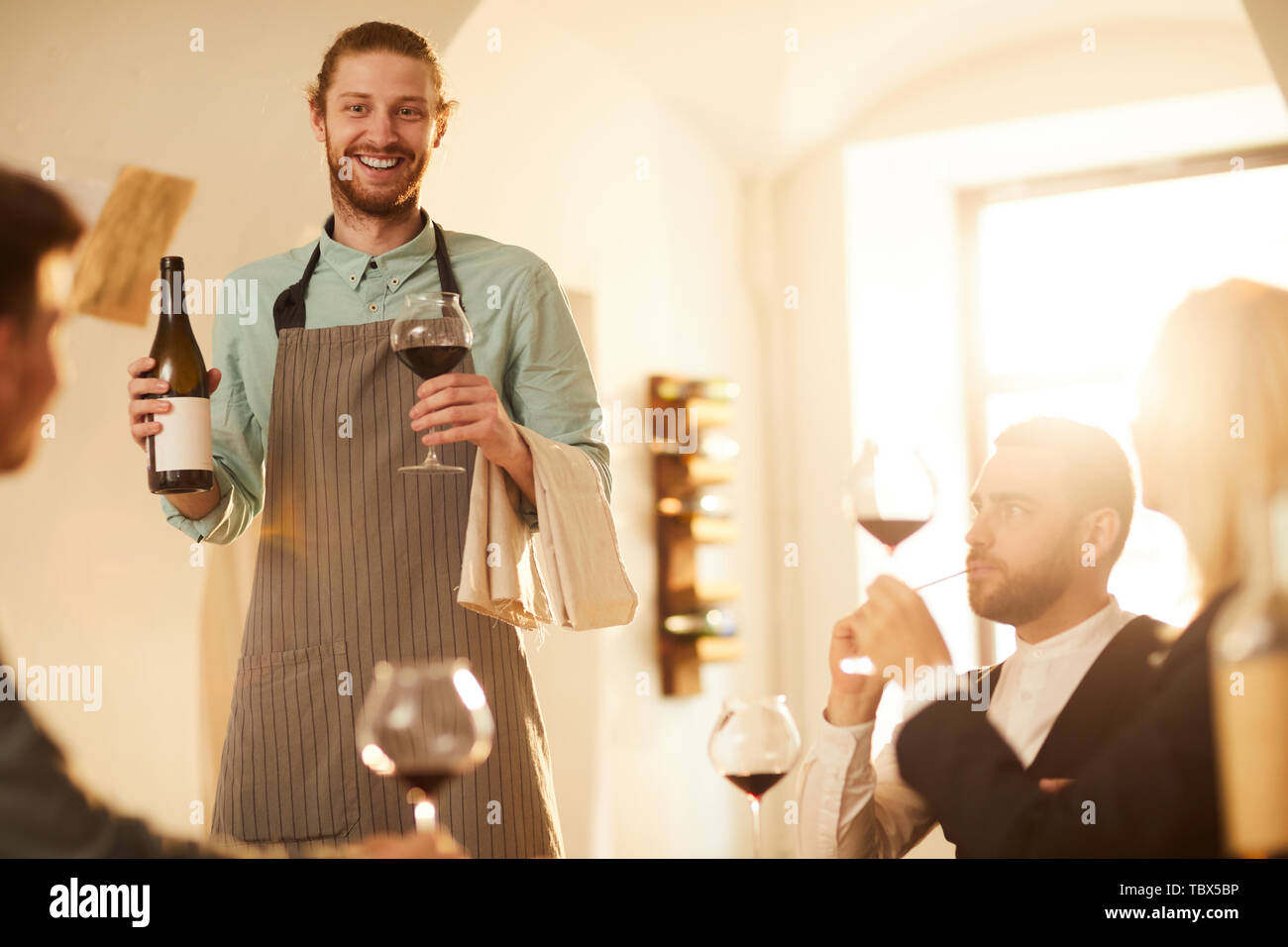 Portrait of cheerful waiter bringing bottle of wine at vineyard restaurant lit by sunlight, copy space Stock Photo