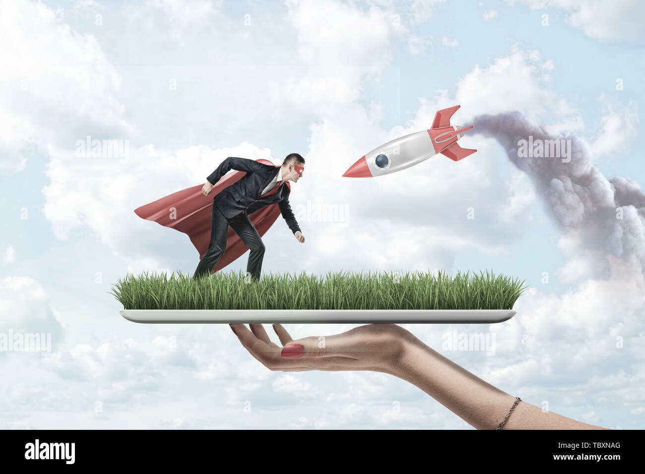Female hand holding grass board with small businessman in red superman mask and cloak with silver red rocket on blue sky background Stock Photo