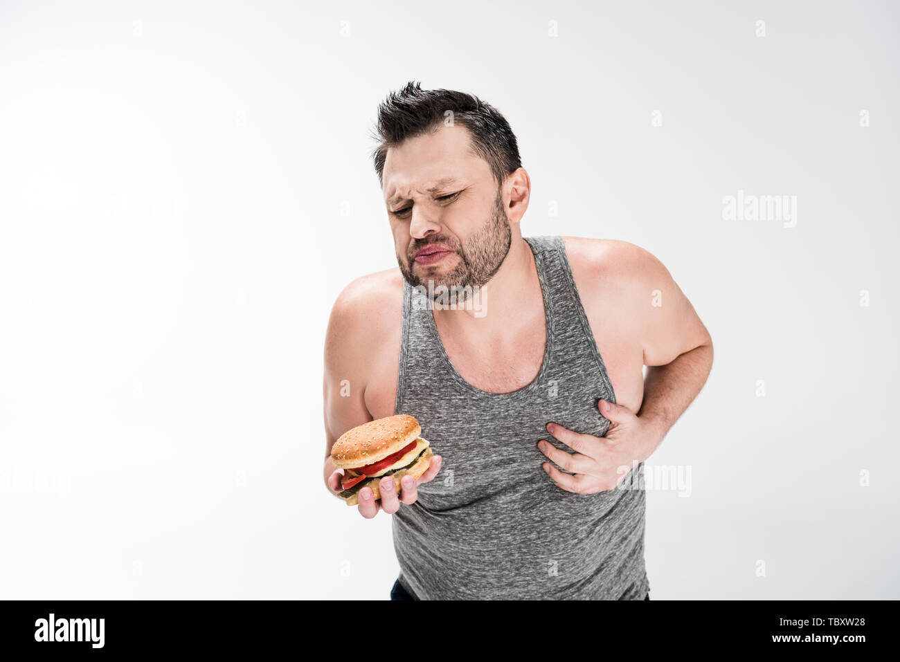 overweight man holding burger and touching chest on white with copy space Stock Photo