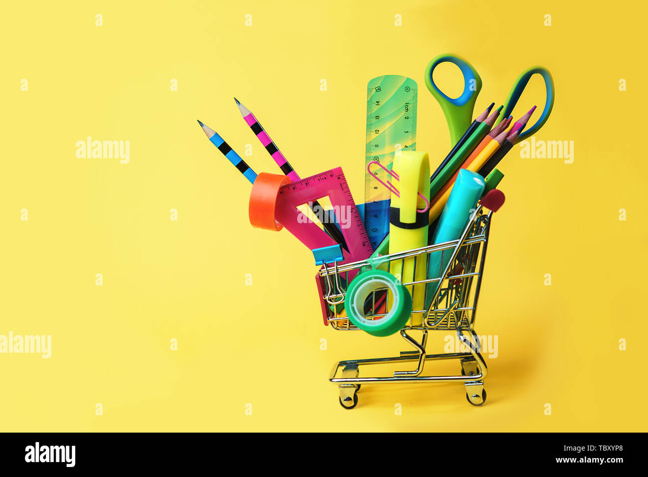 Back to school concept with shopping cart and colorful pencils, square ruler, scissors, clips, markers on pastel yellow backdrop. Stock Photo