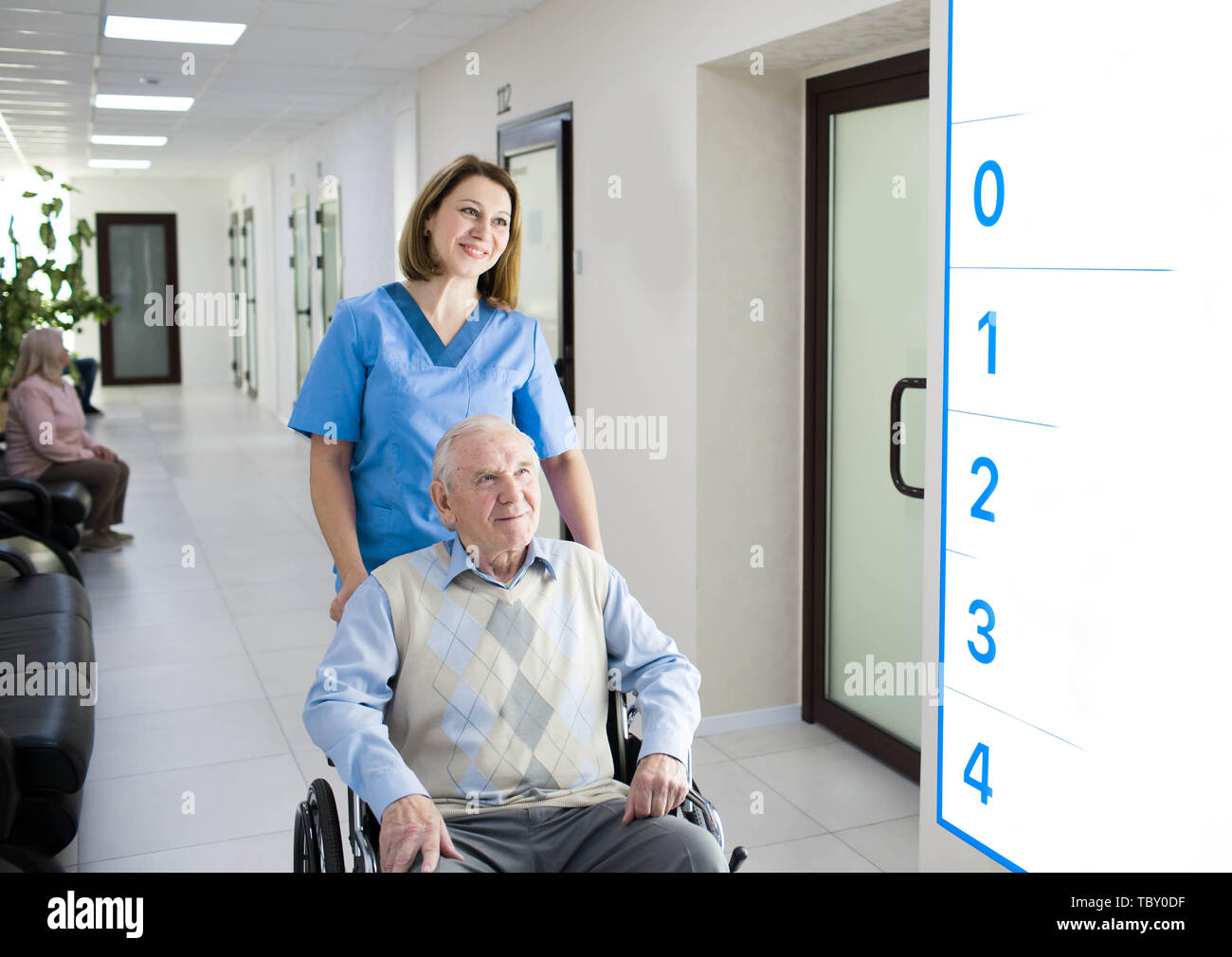Smiling nurse assisting senior man in wheelchair at hospital corridor Stock Photo