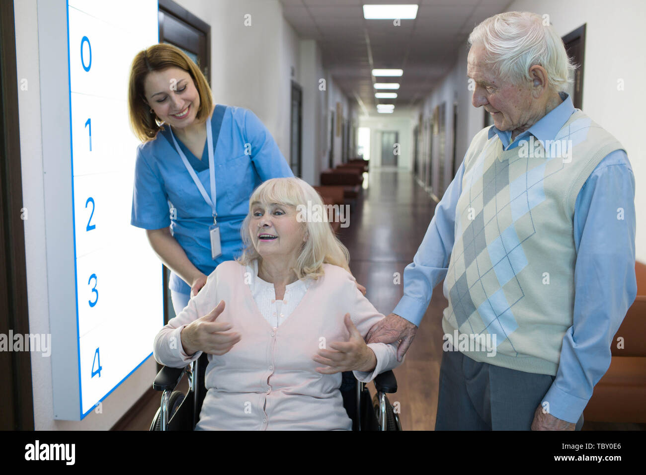 senior woman in wheelchair talking with nurse in a hospital behind her standing senior husband. Stock Photo