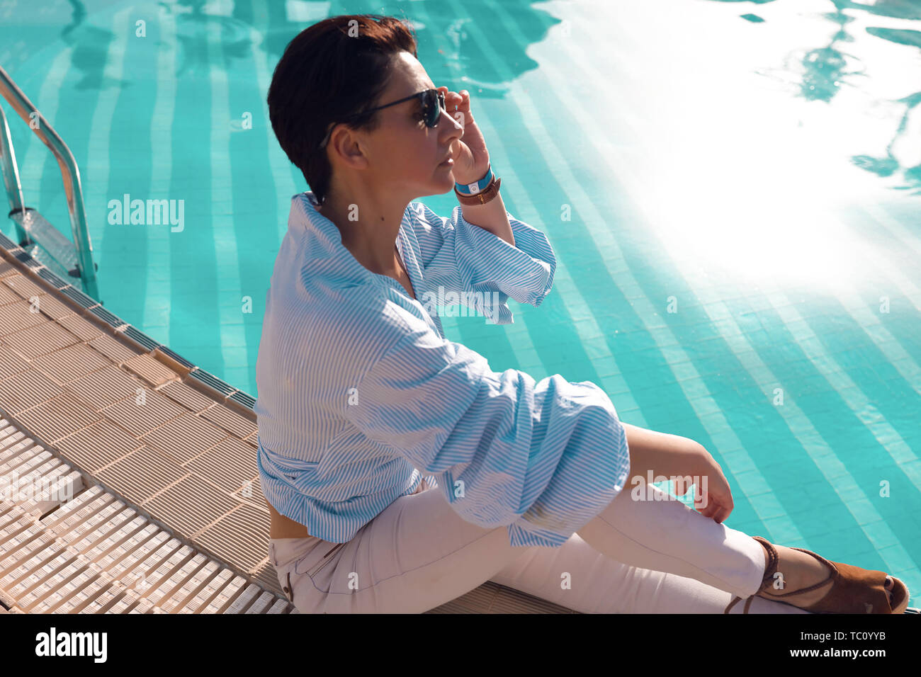Brunette lady sitting near the pool with clear water. Wearing casual clothes, shirt with long sleeves and white jeans. Sunglasses on the face, short Stock Photo