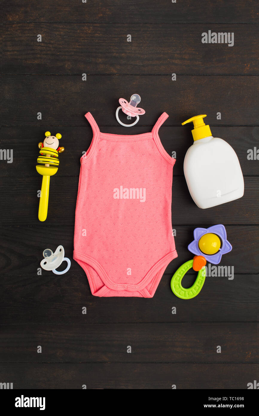 Baby shower accessories. Pink summer bodysuit, two sylicone pacifiers and two wooden and plastic rattles on wooden background with copy space. Vertica Stock Photo