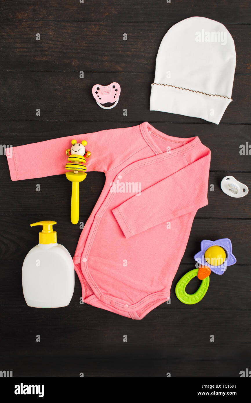 Pink summer bodysuit, hat, children's soap two sylicone pacifiers and two wooden and plastic rattles on brown wooden background with copy space. Baby  Stock Photo