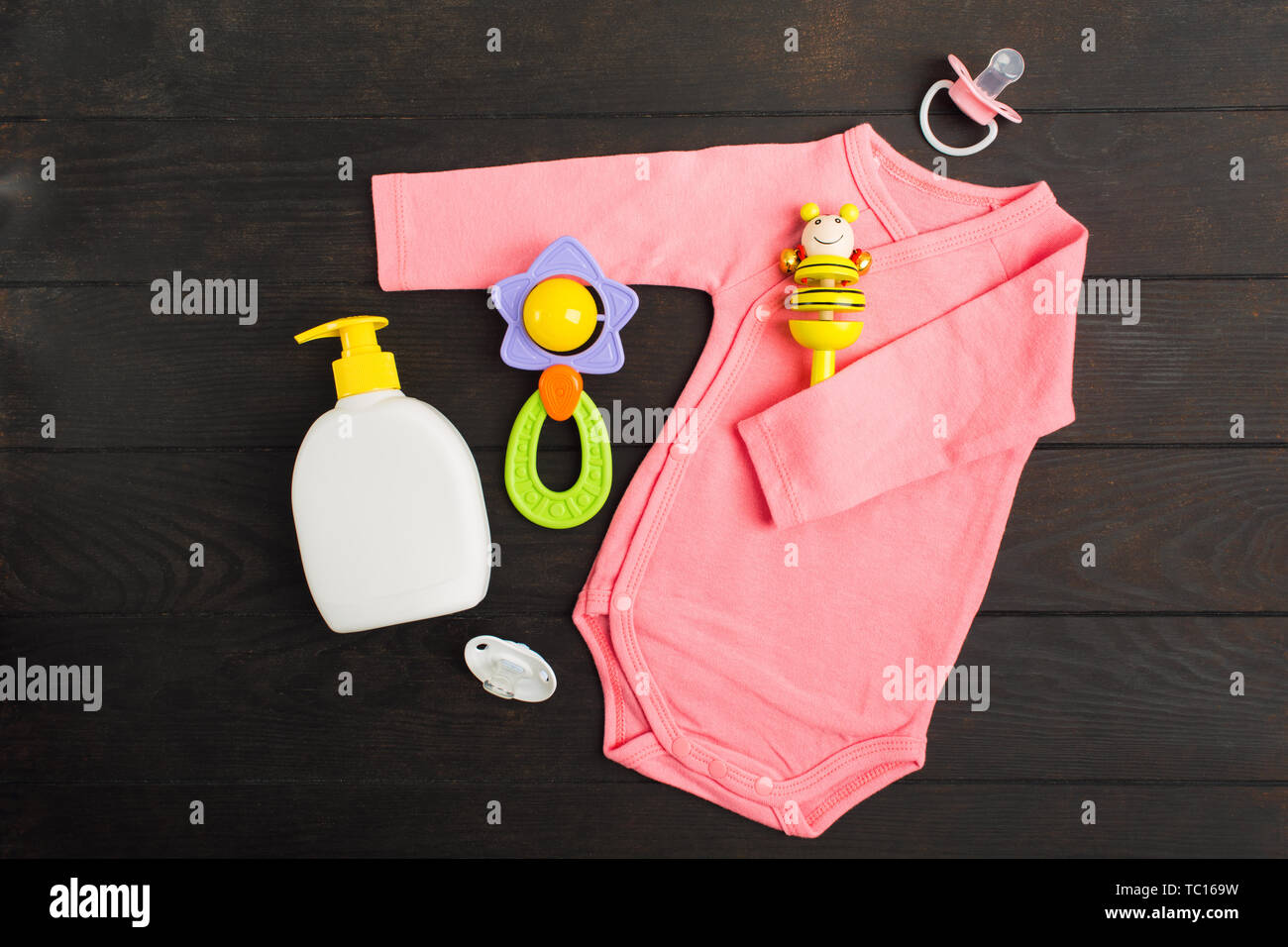 Baby accessories. Flat lay. Pink bodysuit, rattles, baby soap and pacifiers on brown wooden background. Top view Stock Photo