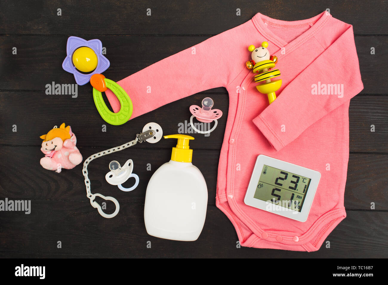 Baby accessories and digital thermo-hygrograph on dark wooden background. Pink bodysuit, two rattles, two pacifiers, toy and soap with hygrometer and  Stock Photo