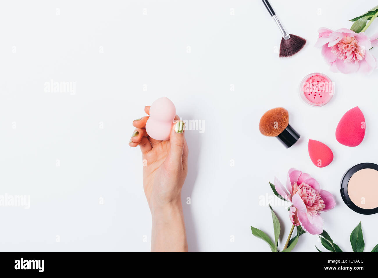 Female hand holding sponge beauty blender next to pink peonies, cosmetic brushes, powder and blush, flat lay on white background. Stock Photo