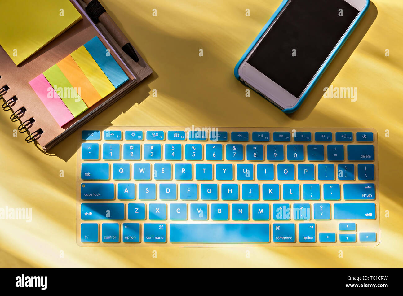 Top view of Workspace with keyboard, smartphone and Spiral Notebook with Pen in Holder with colorful sticky page markers on yellow background Stock Photo