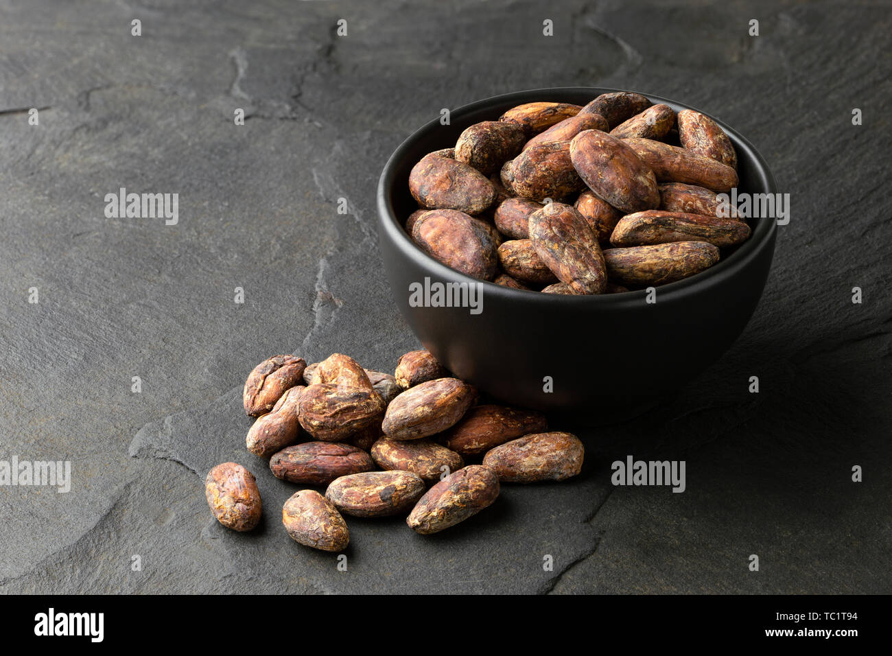 Roasted unpeeled cocoa beans in a black ceramic bowl next to a pile of unpeeled cocoa beans isolated on black slate. Stock Photo