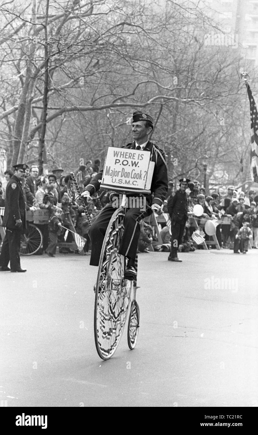 A man wearing a sign reading 'Where is POW Major Don Cook USMC?' rides a penny-farthing or high wheel bicycle, participating in the Vietnam War-related Home With Honor Parade, New York City, New York, March 31, 1973. () Stock Photo
