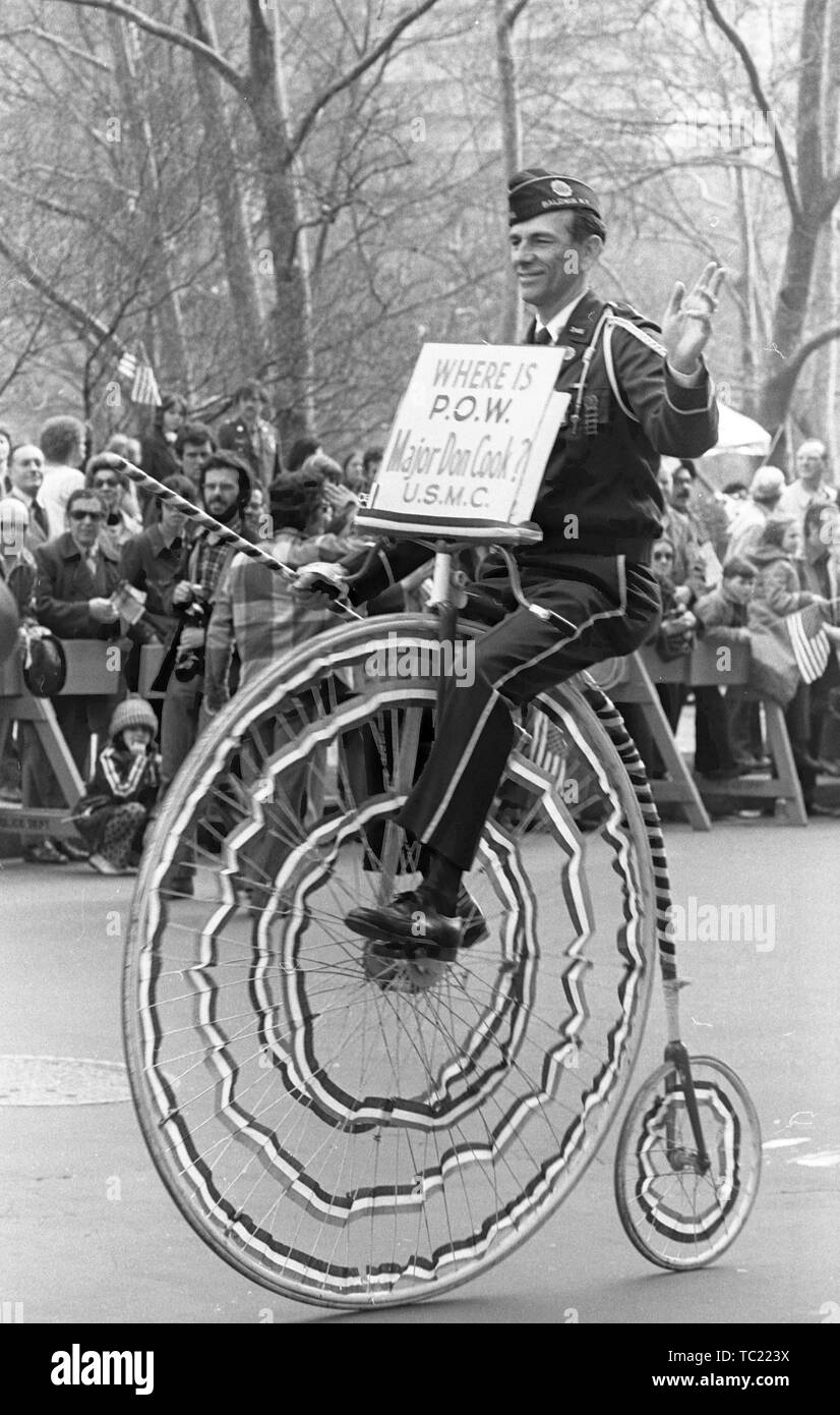 A man wearing a sign reading 'Where is POW Major Don Cook USMC?' rides a penny-farthing or high wheel bicycle, participating in the Vietnam War-related Home With Honor Parade, New York City, New York, March 31, 1973. () Stock Photo