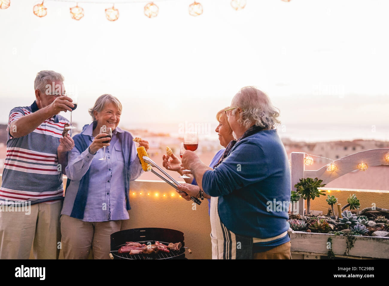 Happy senior friends having doing barbecue party on patio - Retired people having fun drinking wine and grilling meat at bbq dinner at home Stock Photo