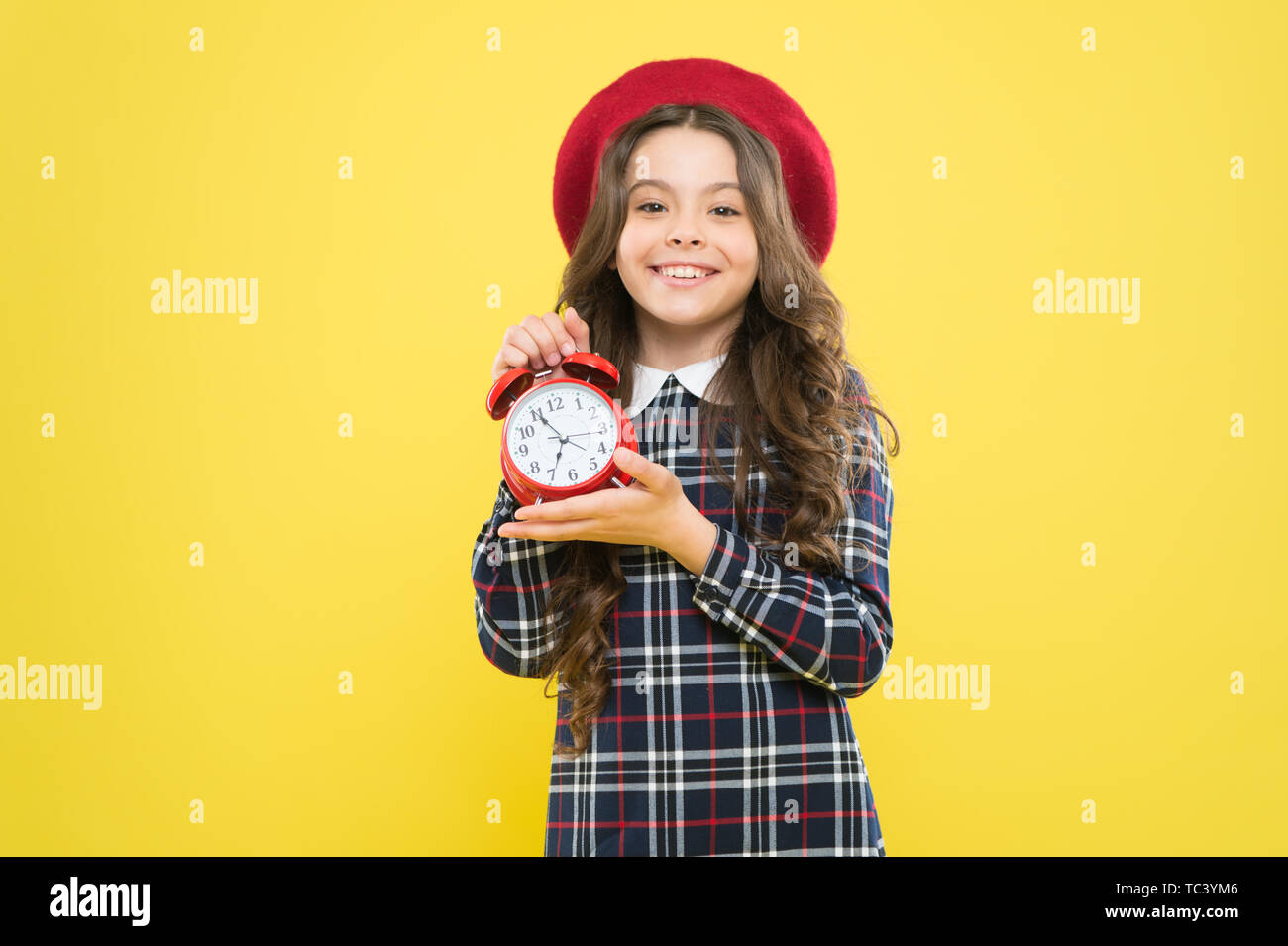 Timing is everything. Happy small child showing proper timing on yellow background. Little girl smiling and holding alarm clock with timing signal. Her timing is perfect. Stock Photo