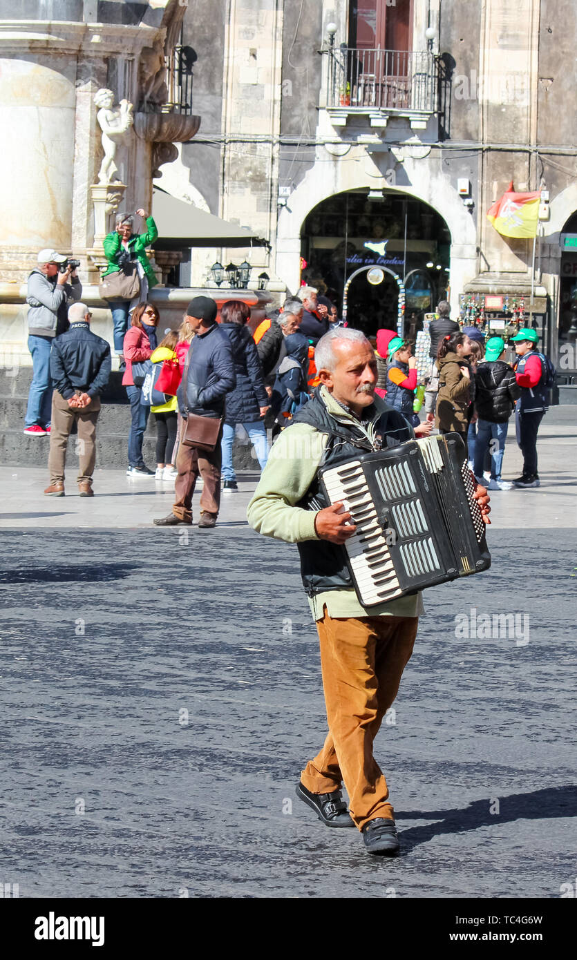 Catania, Sicily, Italy - Apr 10th 2019: Older grey man busker playing accordion on the Piazza Duomo square in the old town. Entertaining street performance for gratuity. Stock Photo