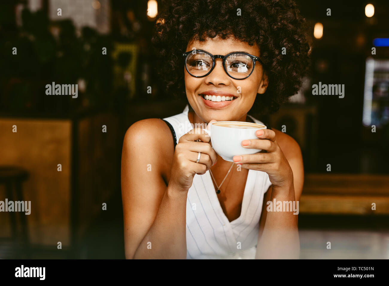 Beautiful young woman sitting at cafe with a cup of coffee and looking away smiling. African female having coffee at coffeeshop. Stock Photo