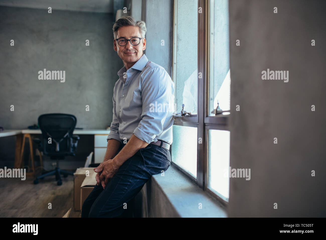 Happy mature businessman leaning by a window sill and staring to camera. Male entrepreneur at his office looking at camera. Stock Photo