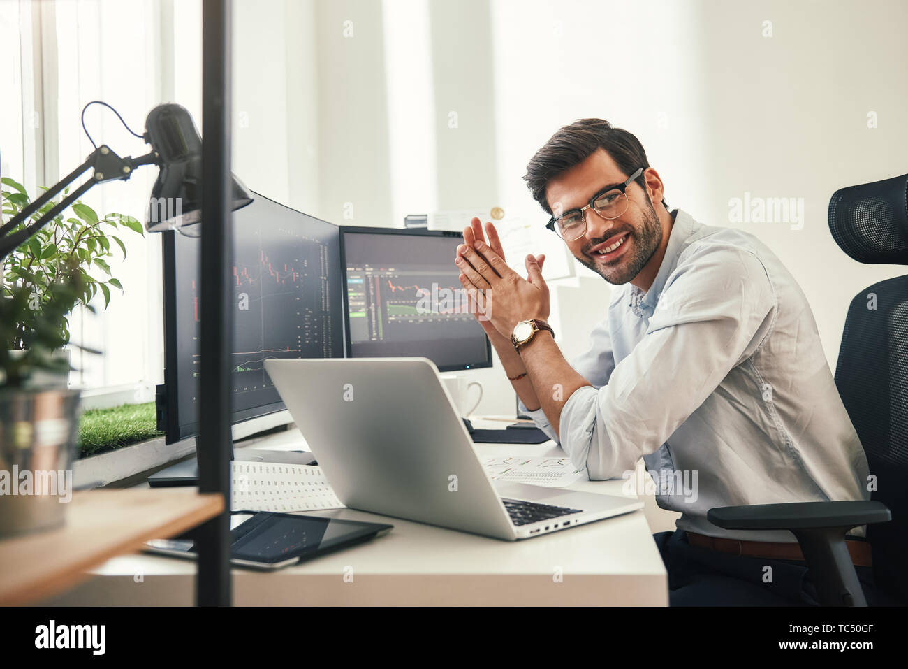 Young and successful. Happy bearded trader in formal wear and eyeglasses looking at camera and smiling while sitting in his modern office. Stock broker. Forex market. Trade concept Stock Photo
