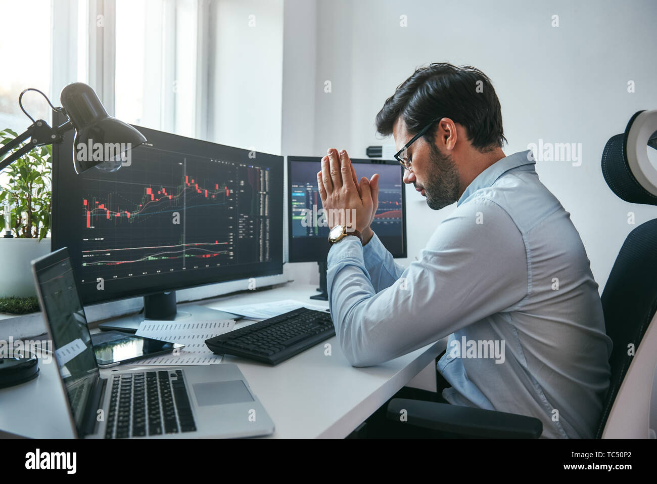 Hoping for the success. Side view of tired young businessman or trader holding palms together while working with data and charts on computers at his modern office. Forex market. Trade concept. Business concept Stock Photo
