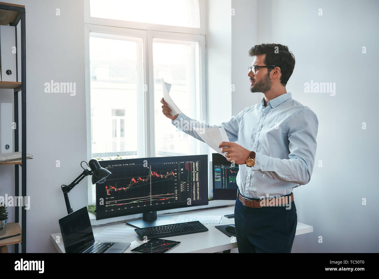 Trading strategy. Smart and young trader in eyeglasses looking at financial reports and analyzing trading charts while standing in front of computer screens in modern office. Stock broker. Forex market. Trade concept Stock Photo