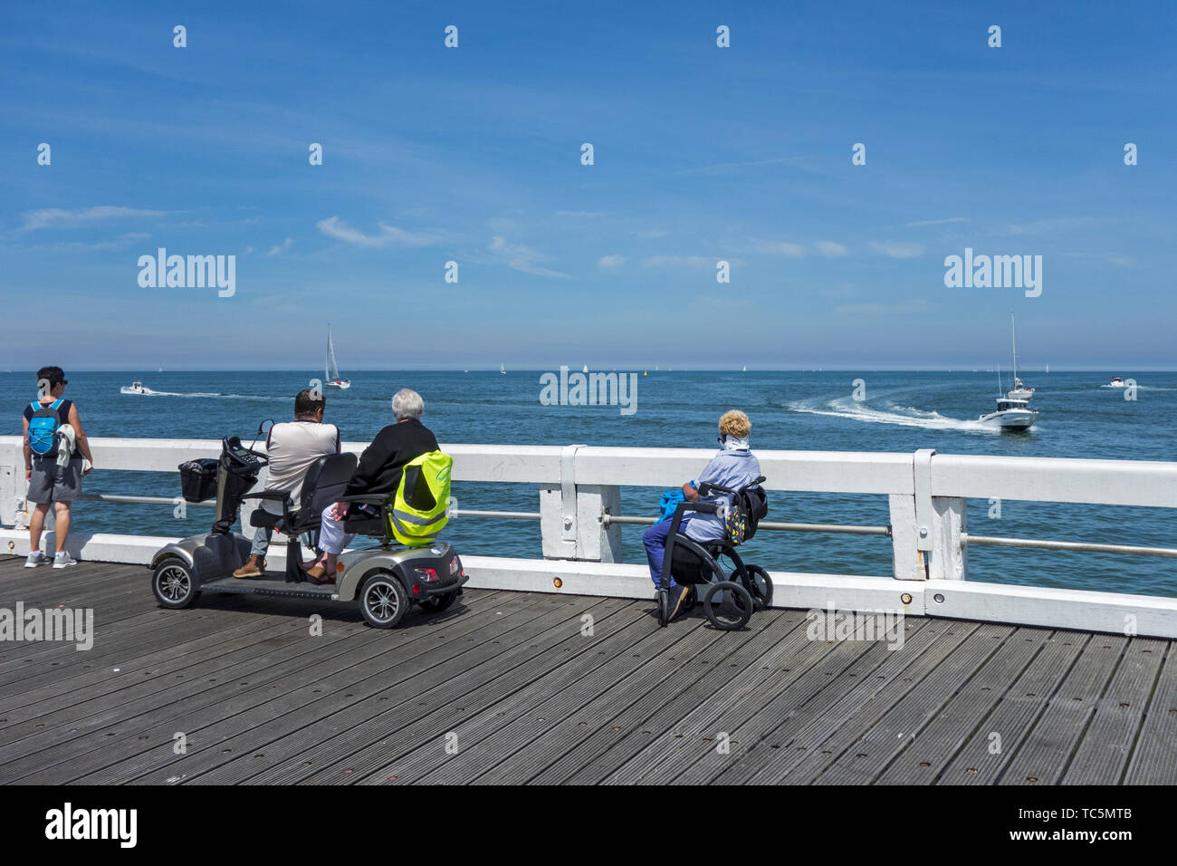 Disabled woman in wheelchair and handicapped persons in duo two person mobility scooter watching boats at sea from jetty Stock Photo