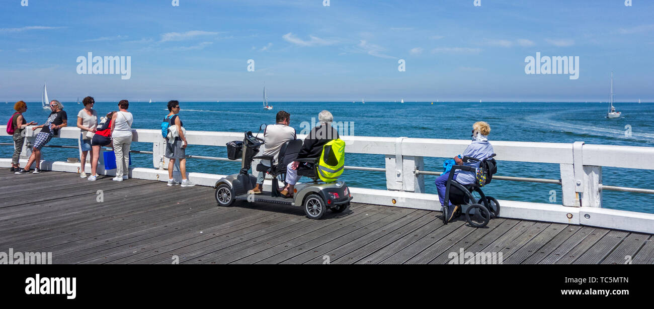 Disabled woman in wheelchair and handicapped persons in duo two person mobility scooter watching sailing boats at sea from jetty Stock Photo