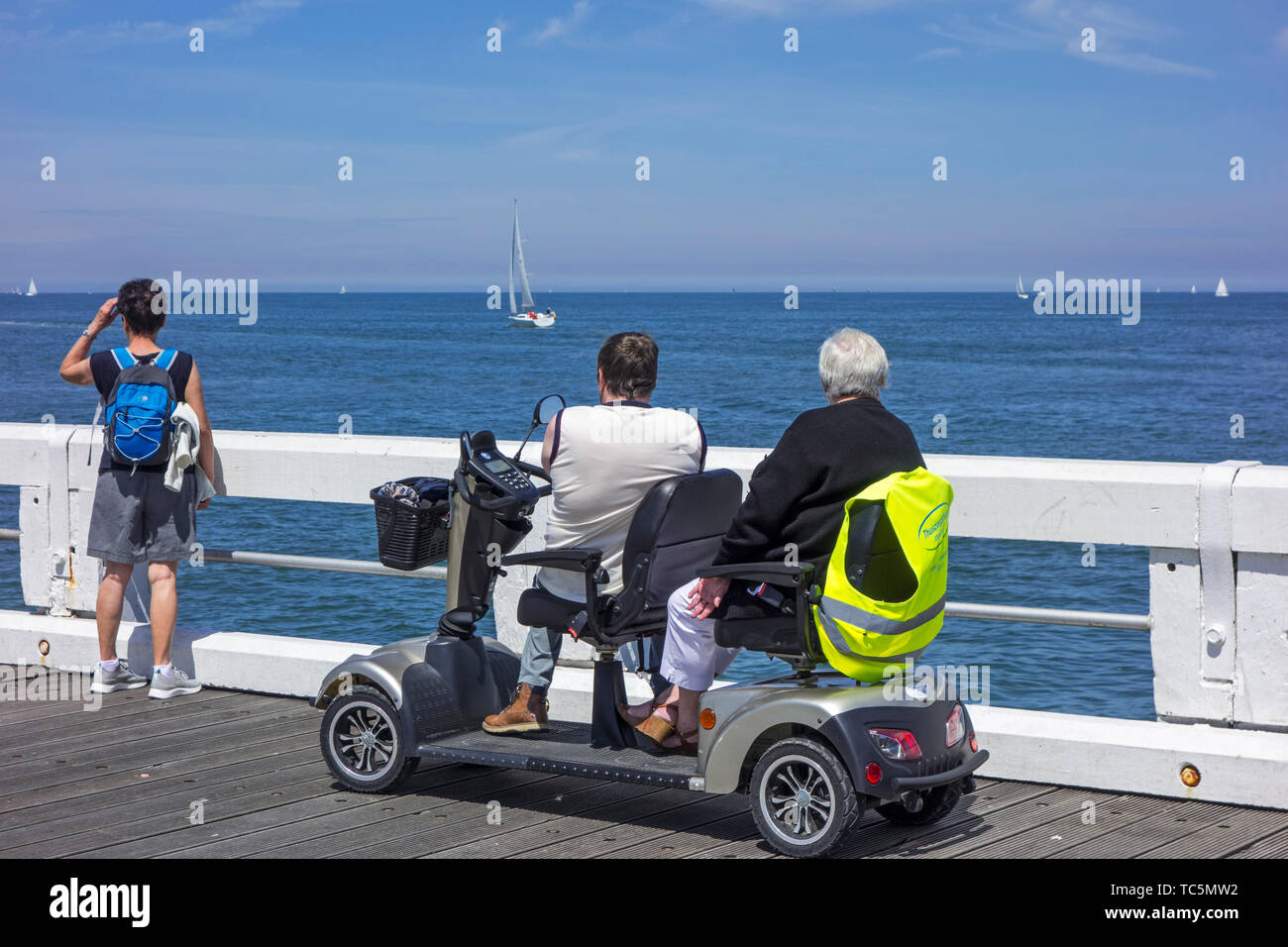 Disabled pensioners in duo two person mobility scooter watching sailing boats at sea from jetty at seaside resort in summer Stock Photo