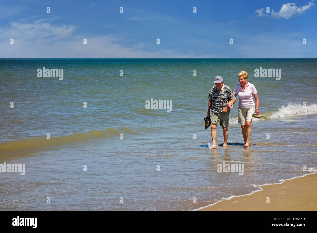 Happy elderly couple paddling in shallow sea water on sandy beach along the coast in summer Stock Photo