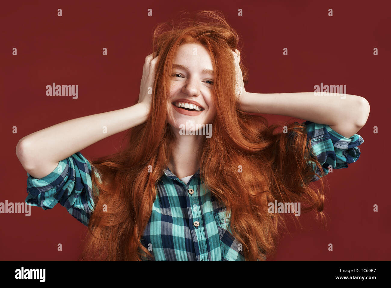 Happy beautiful girl in checkered shirt smiling at the camera while touching her red silky hair over red background. Front view Stock Photo
