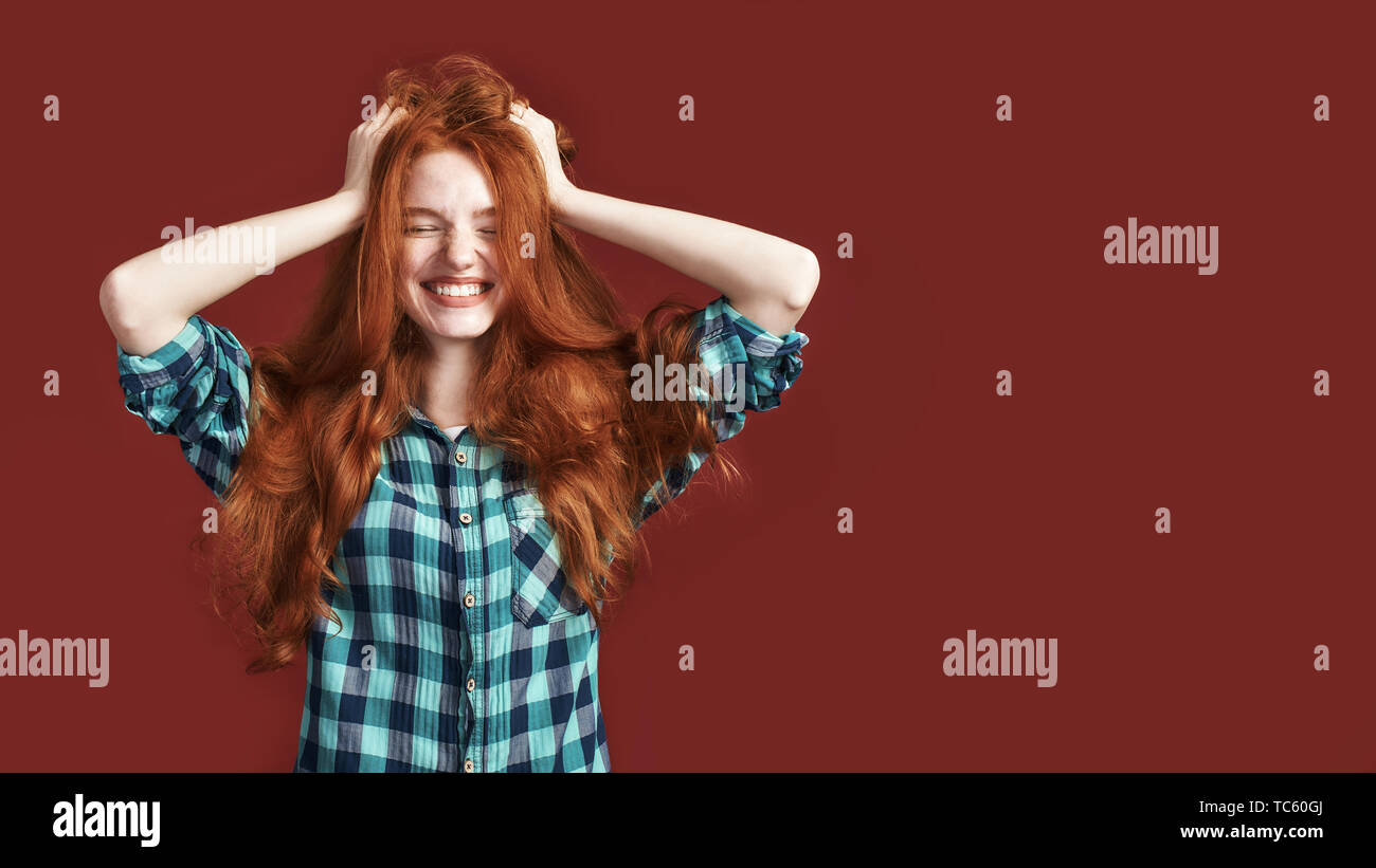 Happy beautiful girl in checkered shirt smiling with closed eyes touching her red silky hair over red background. Front view Stock Photo