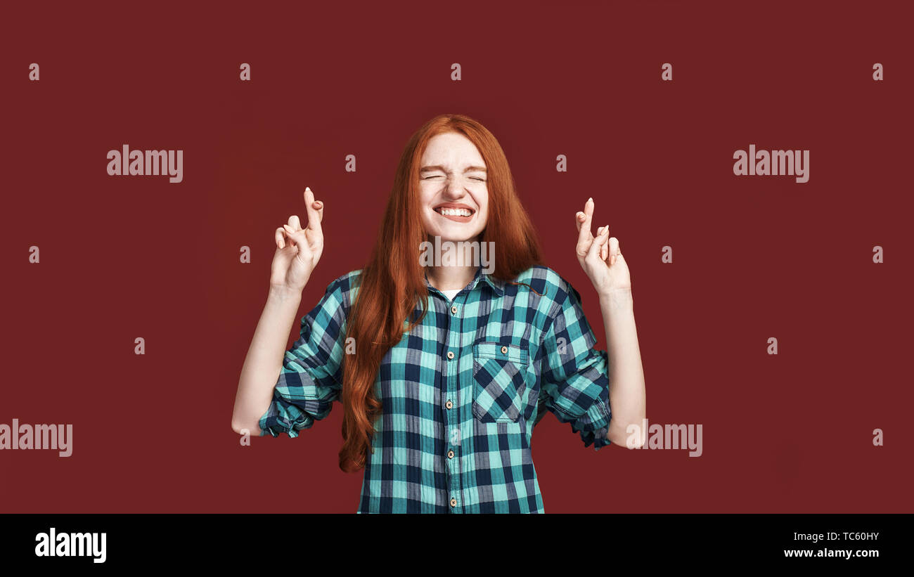 Girl dreams about fulfilling dream, cross fingers for good luck, bite lower lip as eager for wish come true, praying with close eyes and thrilled hopeful expression posing over red background Stock Photo