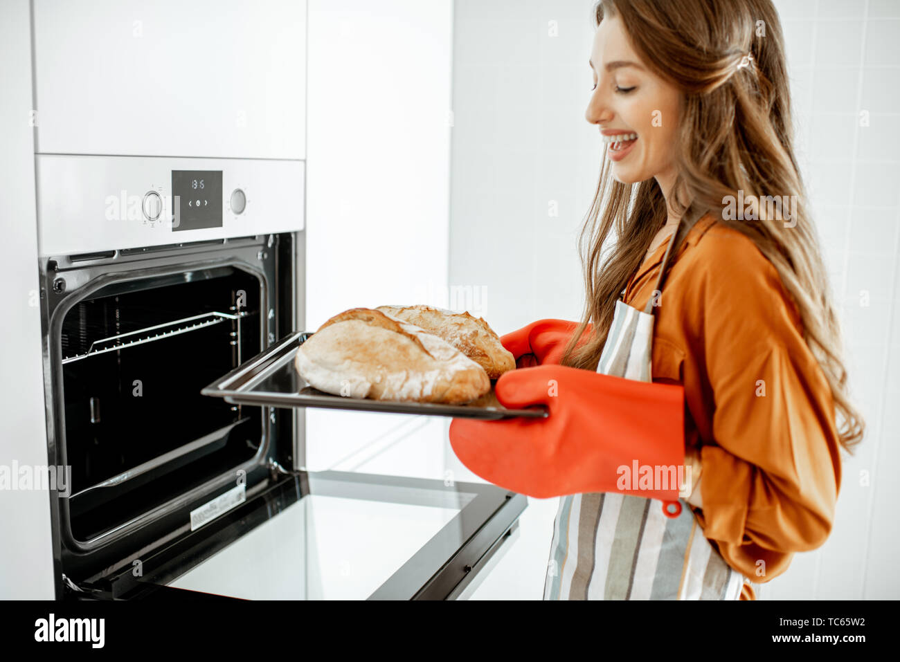 Beautiful young woman baking bread in the oven at the modern white kitchen at home Stock Photo