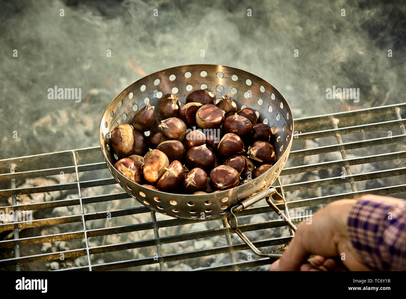 Man roasting a batch of fresh sweet chestnuts on a grill over the hot coals of a barbecue fire in a first person POV Stock Photo