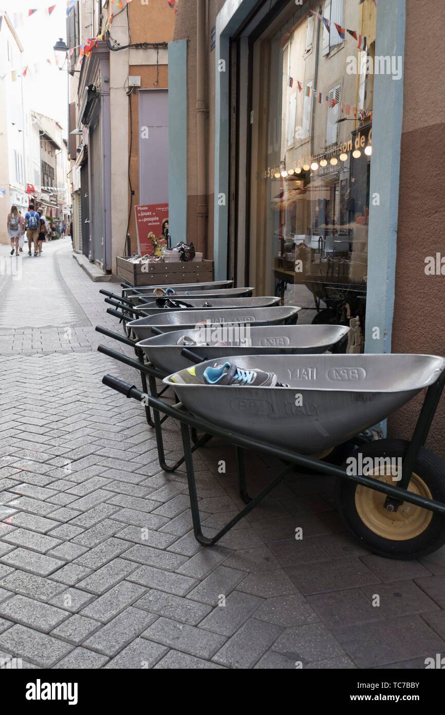Row of horizontal wheelbarrows with shoes and trainers, and ropes of fairy lights and bunting, with tourists in the distance in L'Isle sur la Sorgue, Stock Photo
