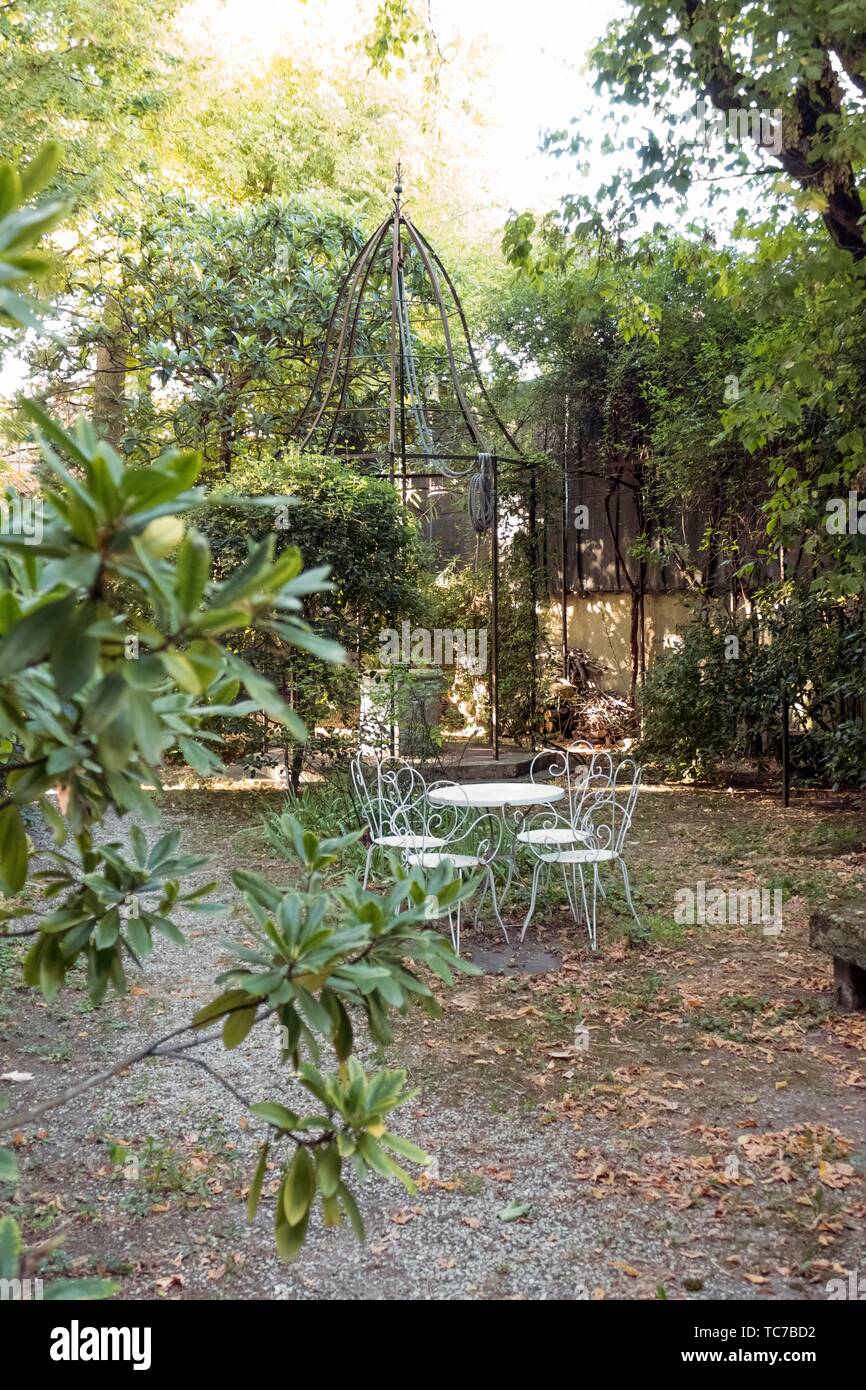 Elegant bell shaped metal canopy protects a stone well in a tree lined courtyard garden with decorative curlicue white cafe table and chairs in Stock Photo